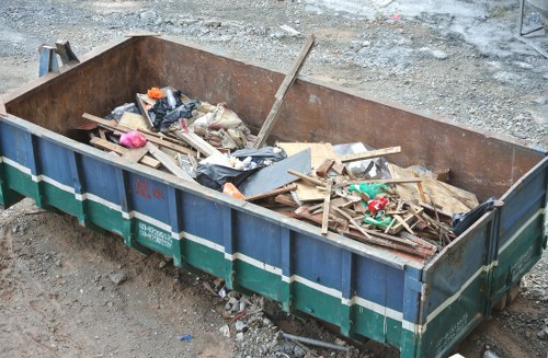 A large, rectangular metal skip with rusted edges and a green and blue exterior, positioned on a gravel surface at a construction or demolition site in Herne Hill. The skip is filled with various types of rubbish including wooden planks, broken pieces of drywall, black plastic bags, and other debris. Some wood panels are stacked loosely, while others are scattered across the top, displaying a mix of textures and finishes such as rough, weathered wood, smooth plastic, and torn packaging. The debris extends to the edges of the skip, with a few items leaning over the sides. The background shows dirt and gravel, typical of a site awaiting clearance or waste removal, with no vegetation or buildings visible. This scene exemplifies typical rubbish disposal at an urban construction site, with a focus on waste material ready for collection by a professional rubbish removal service like Office Clearance Herne Hill.