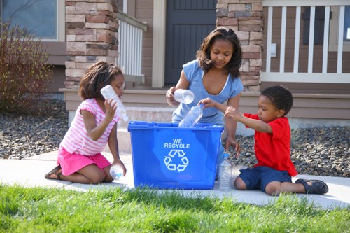 Three children, two girls and one boy, are seated on the grass in front of a residential home in Herne Hill, engaging in a recycling activity with a blue plastic recycling bin that features a white recycling symbol and the words 'WE RECYCLE'. The girl on the left is wearing a pink and white striped top with a pink skirt, holding a clear plastic bottle, while the girl in the middle, dressed in a light blue t-shirt, is placing a plastic bottle into the bin. The boy, wearing a red t-shirt and dark shorts, is handing a plastic bottle to the girl. The background shows the front porch of a house with brick and stone facade, a black door, and white railing, with a gravel garden area to the right. The scene is set during daylight with natural lighting, emphasizing the environmental theme relevant to rubbish and recycling services in the local area.