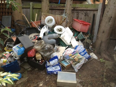 A large pile of mixed household waste and rubbish is accumulated on a patch of bare earth in a backyard or garden area, enclosed by a wooden fence and supported by wooden posts. The debris includes several white plastic pipes with visible dirt, a red plastic storage box or container, crumpled black plastic bags, cardboard boxes, and various smaller items such as food packaging, plastic bottles, and miscellaneous household waste. Items are haphazardly stacked and spilling onto the ground, with some objects leaning against each other or the fence. The background shows tall wooden fence panels and tree trunks, with some foliage visible. The overall scene appears to be awaiting rubbish removal services, possibly from a local provider like Office Clearance Herne Hill, and represents typical garden waste or household clutter collected for disposal or clearance in south London.
