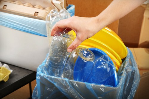A close-up view of a person's hand gripping a clear plastic water bottle, which is being placed into a large blue recycling bag lined with transparent plastic. Inside the bag, there are yellow and blue plastic buckets and containers with textured, ribbed surfaces and glossy finishes. The scene appears to be set indoors, possibly in an office or storage area, with a black surface and a white and blue container visible on the left side. The surrounding environment suggests an organized process of disposing of recyclable plastic waste, aligning with rubbish removal services offered in the Herne Hill area. The materials in the image are mainly lightweight and smooth, highlighting clear plastic and brightly coloured containers, indicating an emphasis on recycling and responsible waste management. The lighting is natural or soft artificial, illuminating the objects evenly and emphasizing the cleanliness and orderliness of the waste sorting activity conducted by Office Clearance Herne Hill.