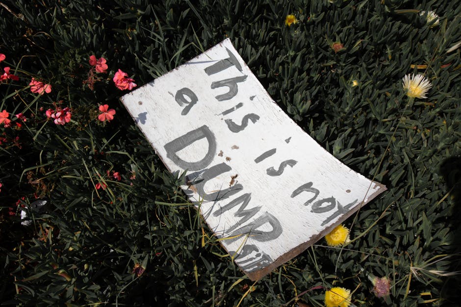 An aerial view of a small excavated area near a dirt pathway, filled with a mound of mixed domestic waste materials. The pile contains crumpled plastic bags, discarded food packaging, paper, and other