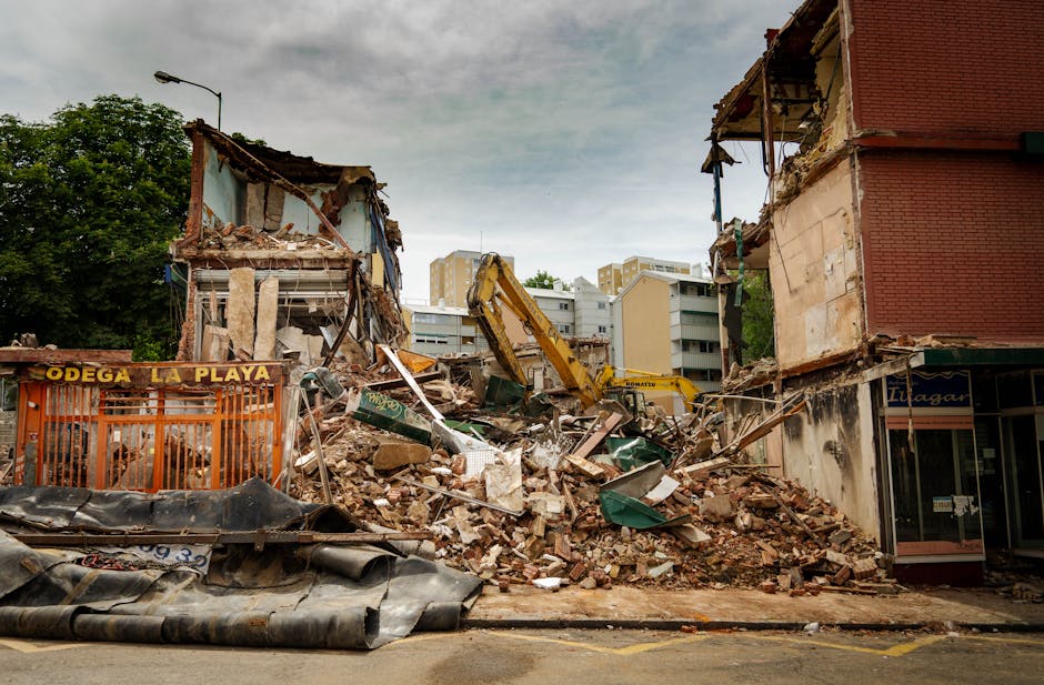 The image depicts a partially demolished brick wall with a white painted surface, exposing uneven brickwork and rubble at the base, including broken bricks, debris, and wooden planks scattered on the 