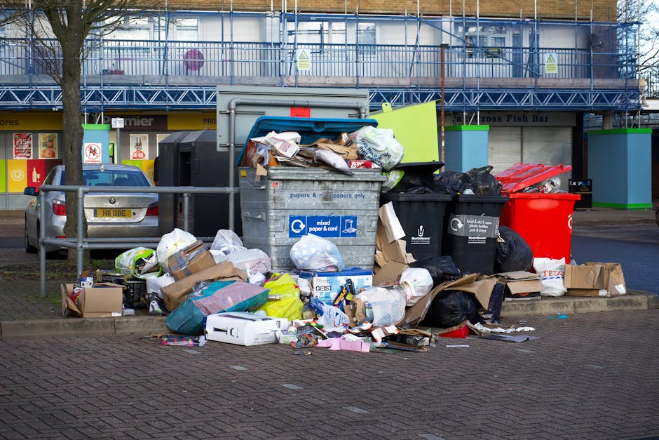 An outdoor scene showing a large pile of mixed waste and rubbish surrounding a white, closed-moving container with a visible registration number, adjacent to a debris-strewn ground. The waste includes